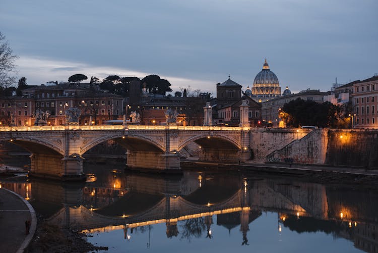 Old Urban Bridge Reflecting In River In Evening City