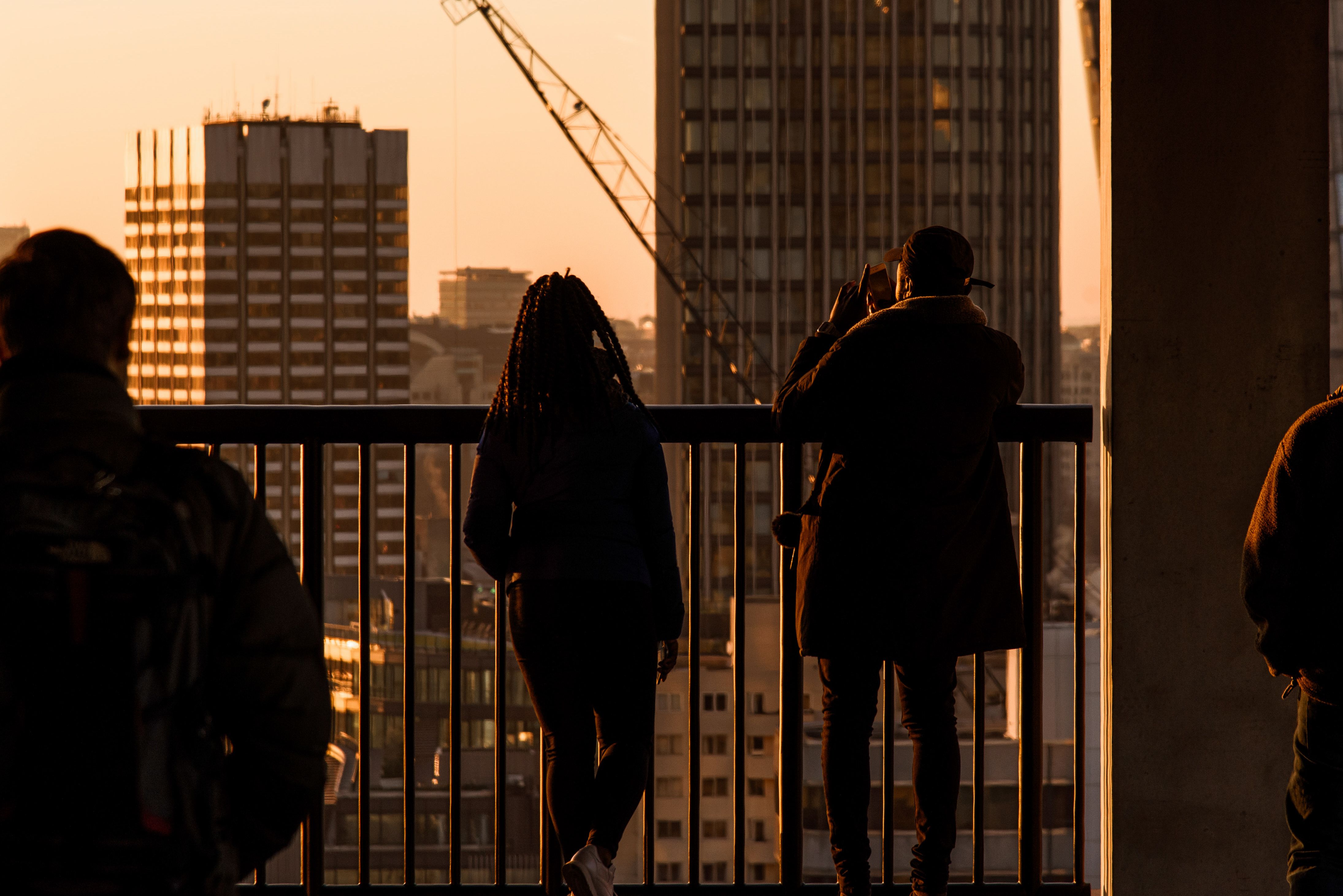 Back view group of unrecognizable travelers standing near metal fence while observing modern multistory buildings located in city in evening time