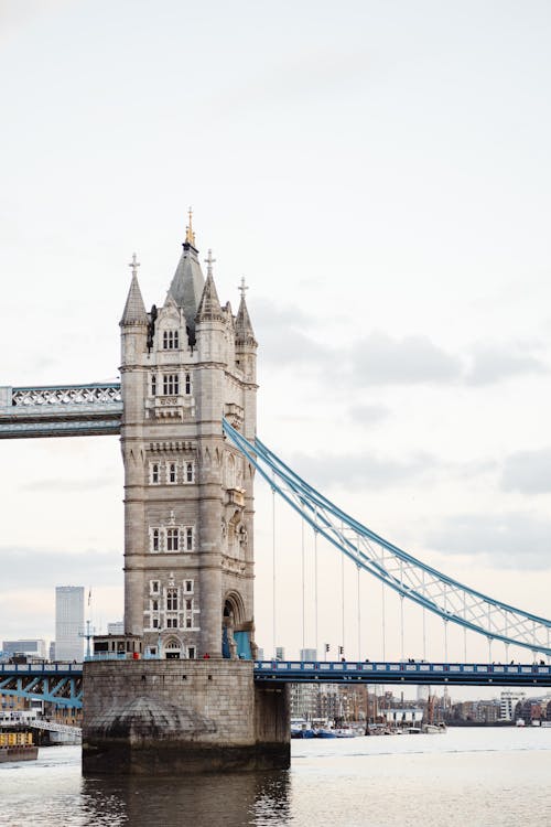 Fragment of famous suspension Tower Bridge crossing Thames river in London city against cloudy sky and shore with residential buildings