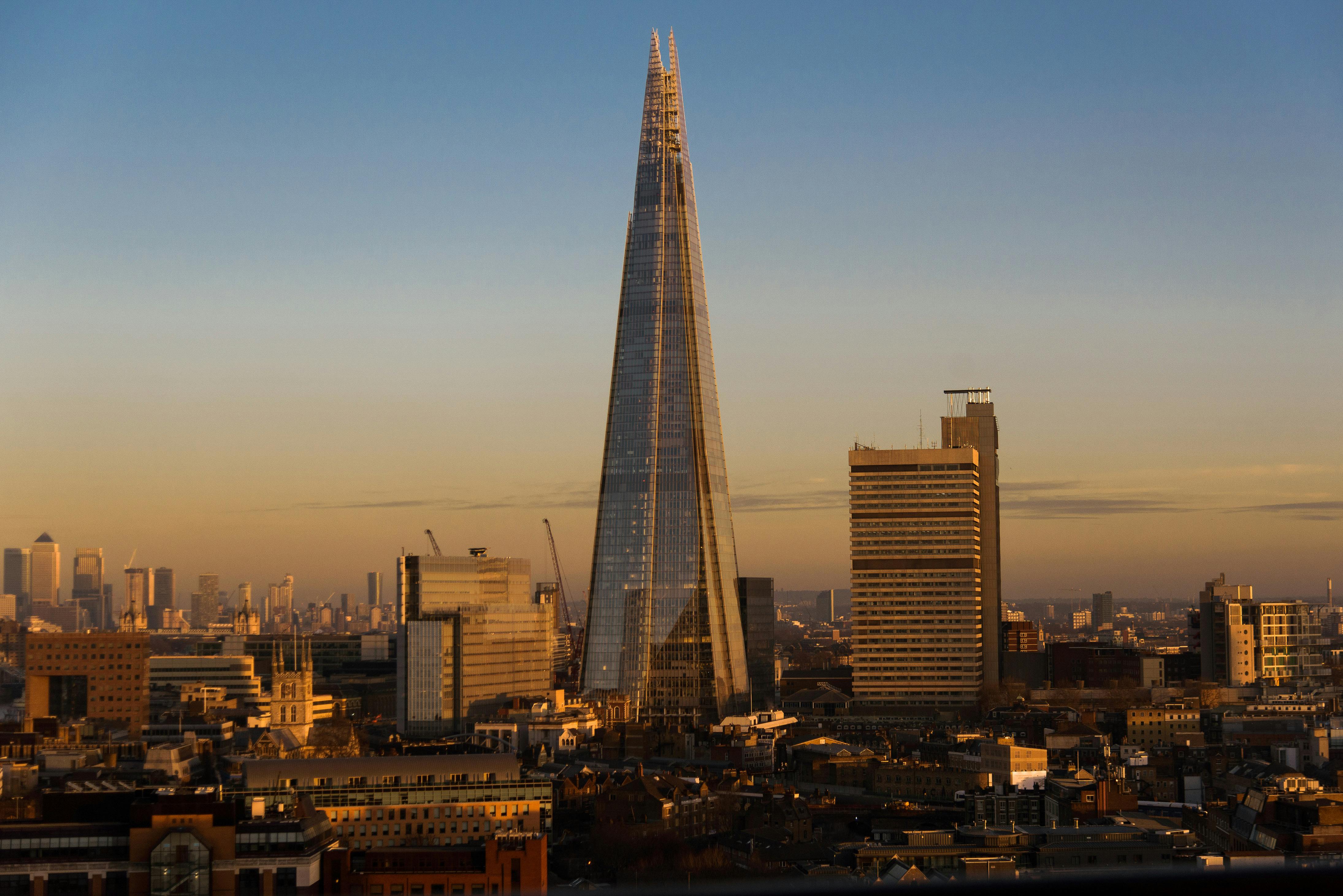 Modern futuristic skyscraper with creative design located on streets with residential buildings in London city in evening time against cloudless sky by Olga Lioncat