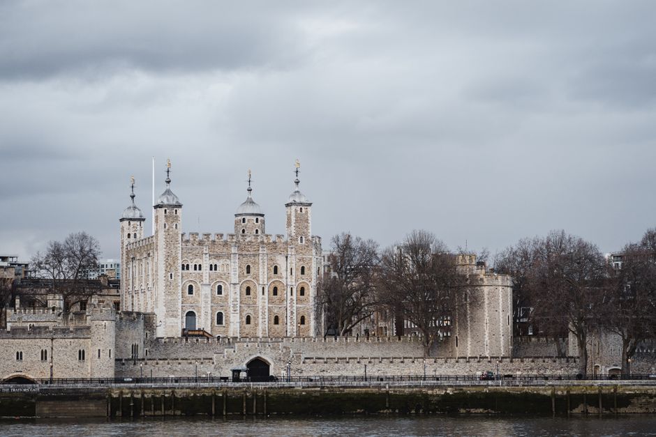 Historic stone White Tower located on river bank near calm Thames against cloudy sky in London city with leafless trees