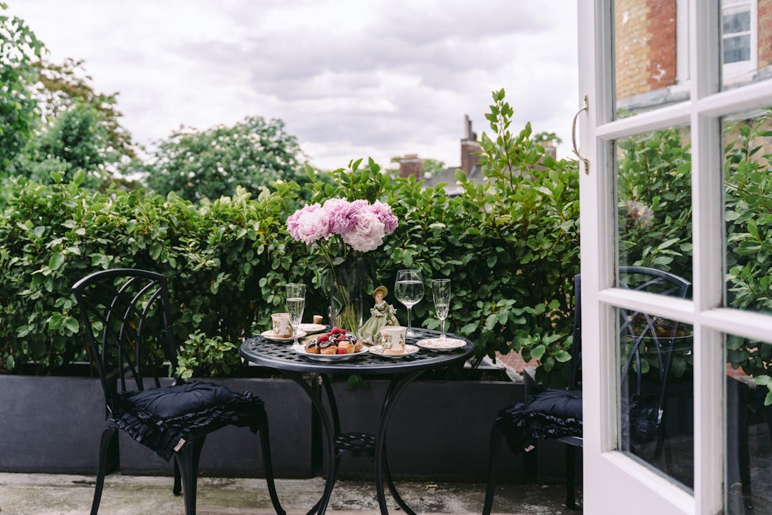Round table with bouquet of fresh gentle peonies in vase composed with wineglasses and coffee cups on terrace of classic styled mansion under cloudy sky