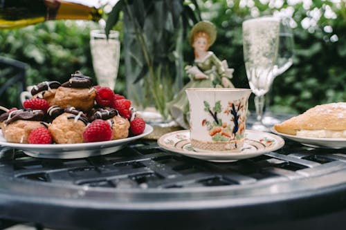 Ornamental porcelain coffee cup and plate with appetizing profiteroles with chocolate and berries served on table with glasses of champagne in outdoor cafe