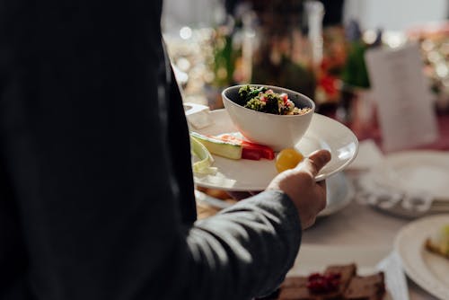 Crop faceless male with bowl of bulgur with spinach and pomegranate seeds on white plate with cut vegetables standing neat table on blurred background