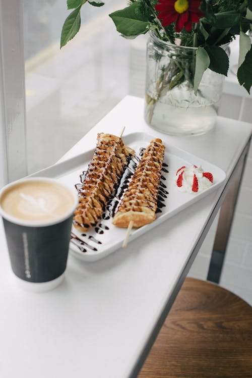 High angle of appetizing fresh waffles with chocolate sauce and paper cup of coffee to go on white table with glass vase