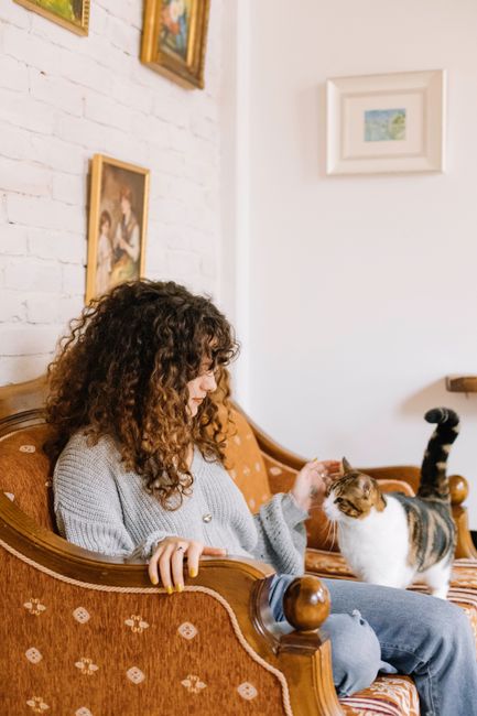 A young woman pets her cat while sitting on a vintage sofa in a warm, stylish living room.