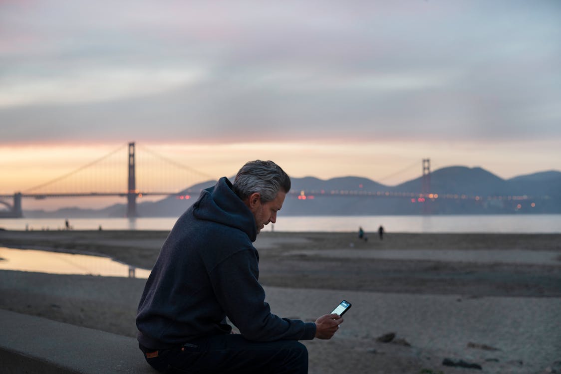 A Man Wearing Jacket Sitting Near the Beach While Using a Smartphone