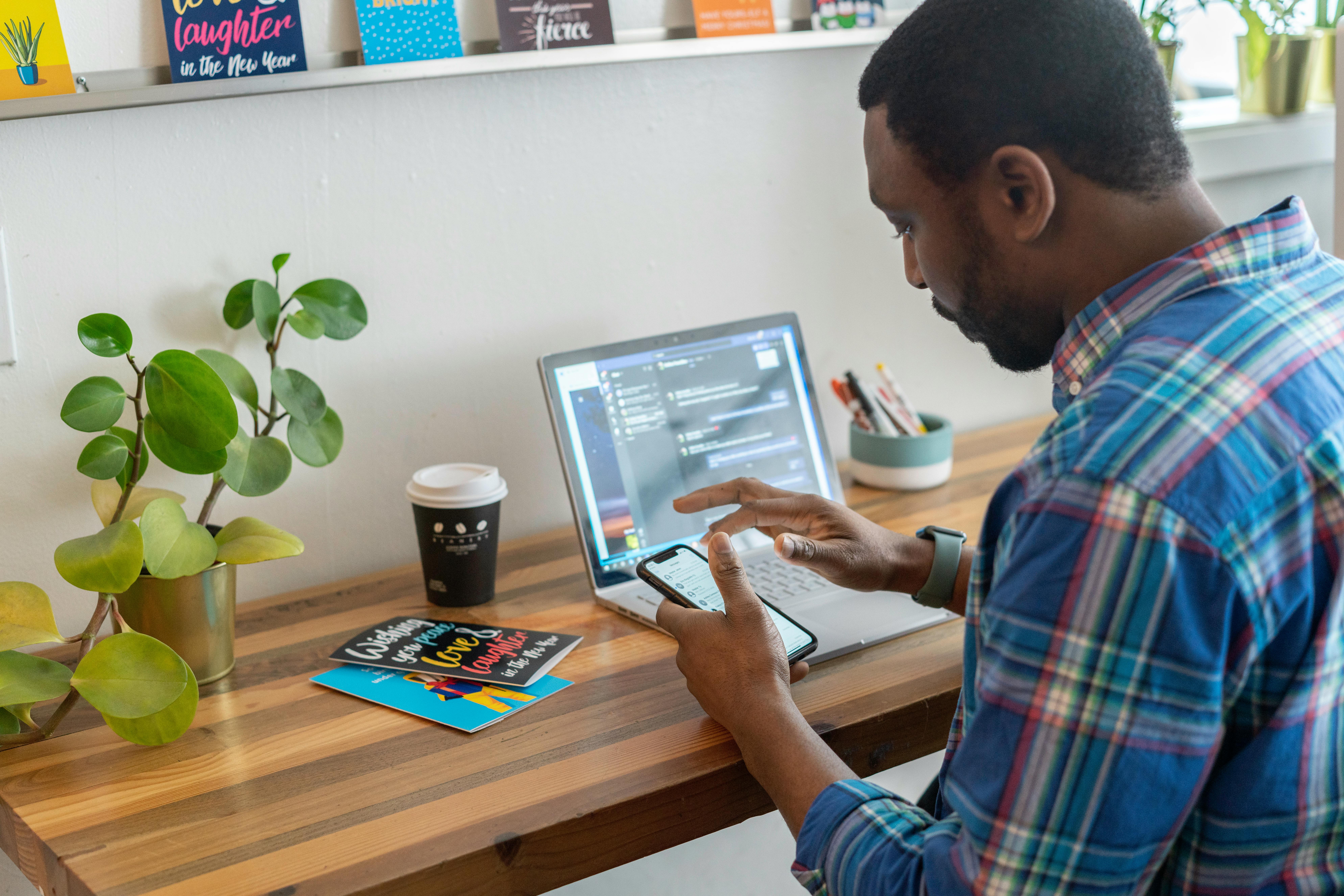 A man uses his smartphone and laptop simultaneously at a wooden desk.
