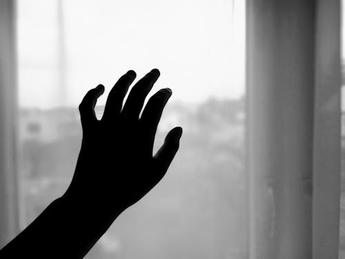 Black and white of crop anonymous lonely person touching window at home in daylight