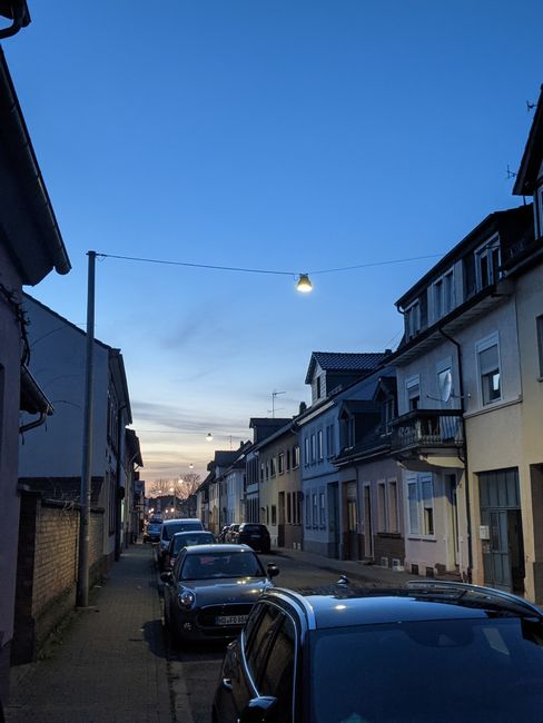 Many automobiles parked on roadside near paved sidewalk between old fashioned residential buildings on street of town in evening time