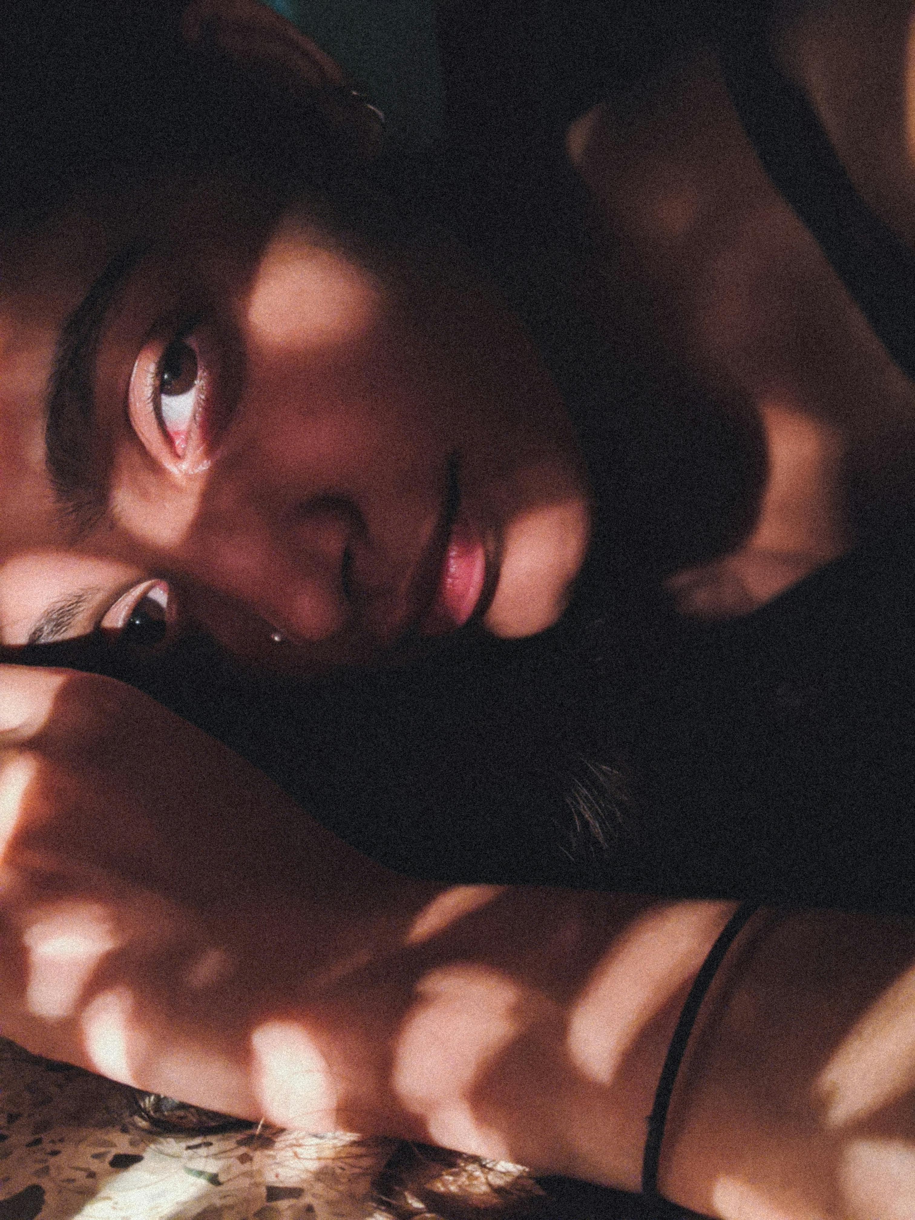 Charming enigmatic ethnic female with pierced nose looking at camera with sincere gaze while lying in obscure room with bright sunlight and shadow