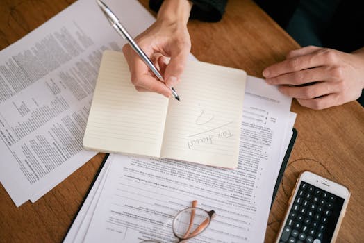 Close-up of hands writing on a notebook surrounded by tax documents and a smartphone.