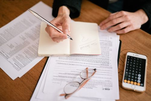 Free Close-up of a person writing in a notebook with documents and a calculator, managing finances. Stock Photo