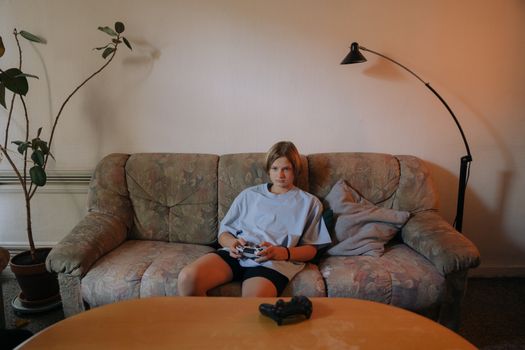 Young teenager sitting on a sofa, playing video games indoors with a controller, surrounded by cozy decor.