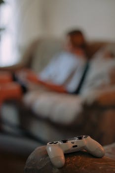 Close-up of a game controller on a sofa with a blurred person in the background, creating a relaxed gaming atmosphere.