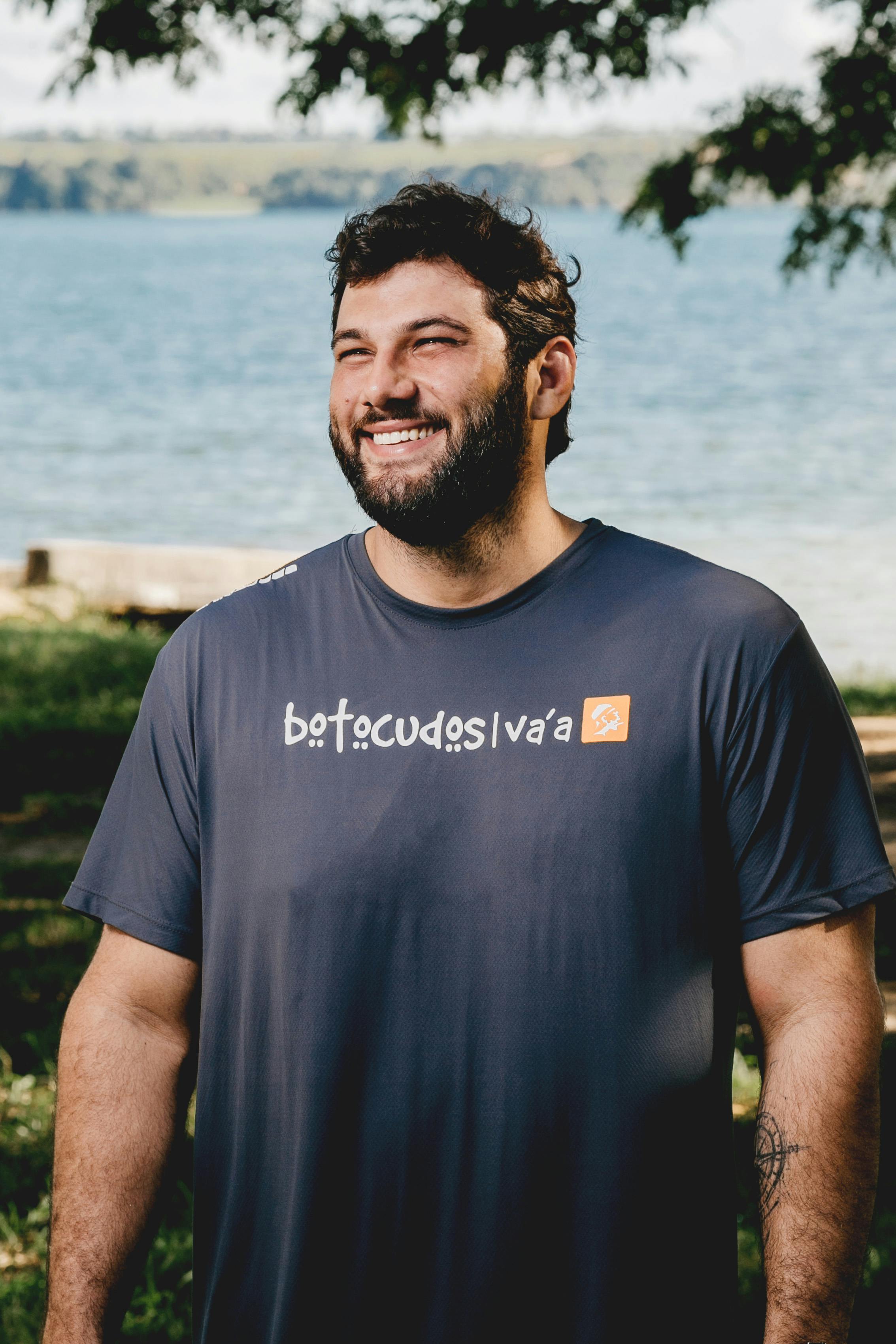 Positive bearded man in t shirt standing on glade against tree branches and blue pond