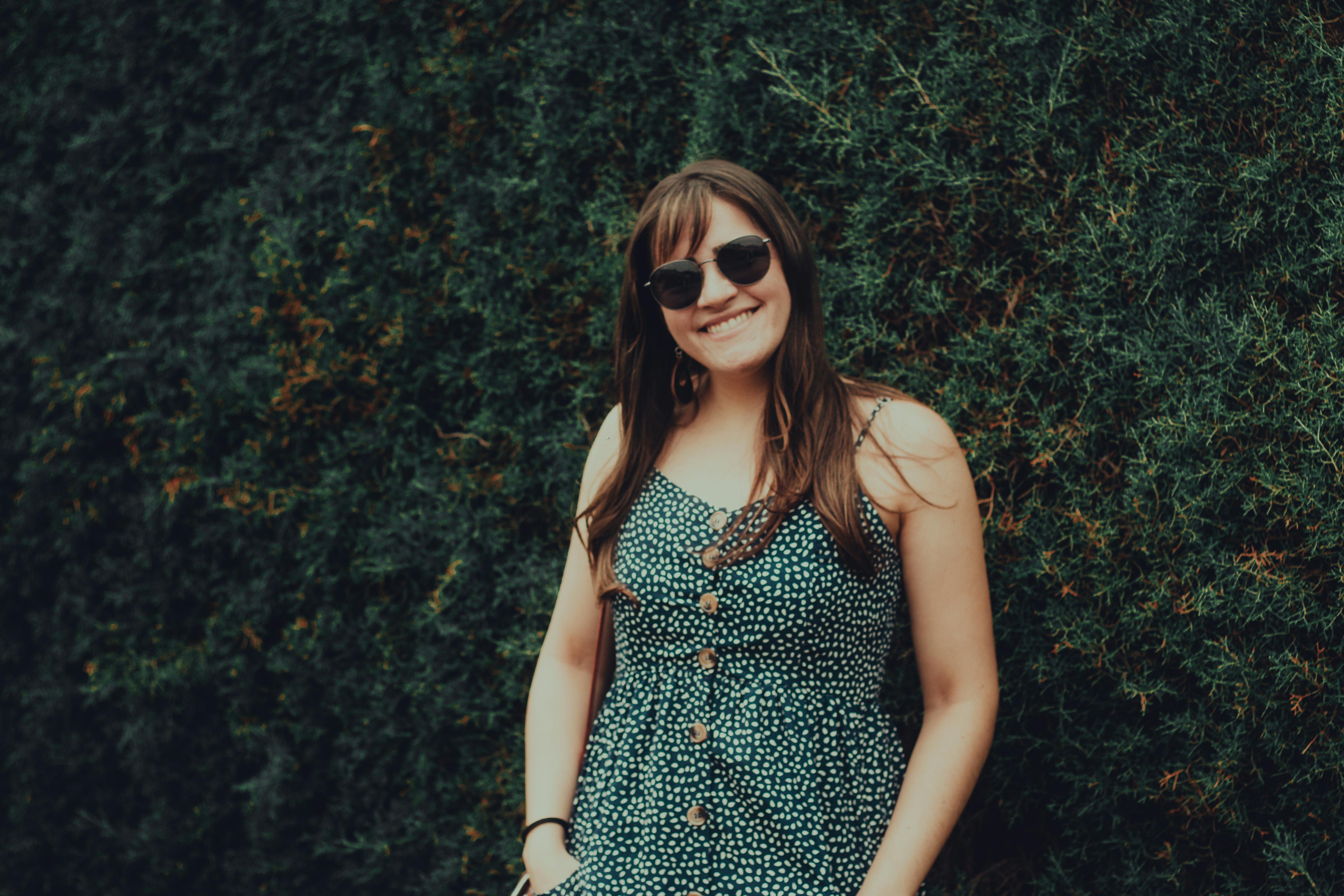Young woman smiling outdoors in a fashionable dress and sunglasses against a lush green backdrop.