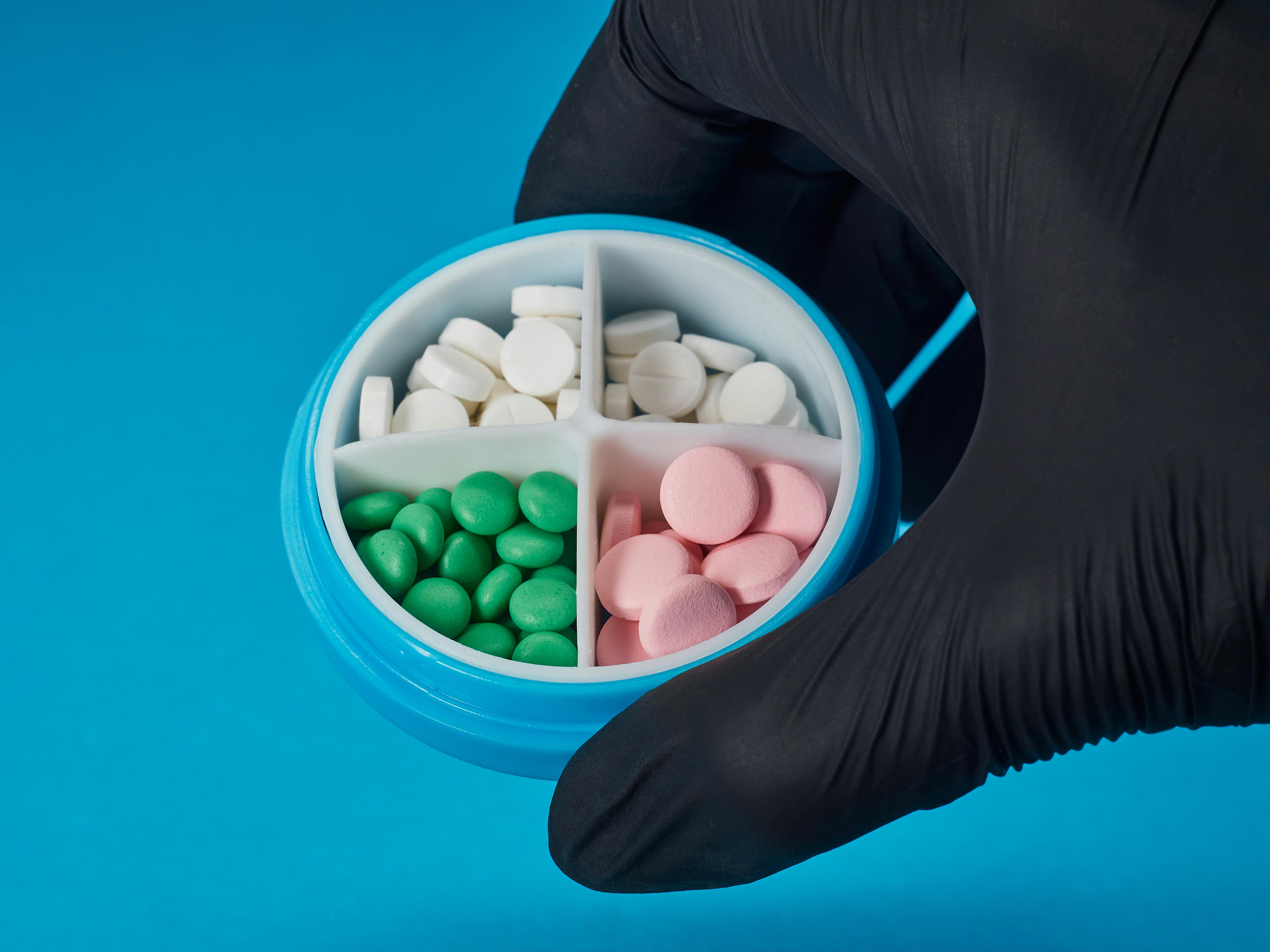 Gloved hand holding a round container with colorful pills on a vibrant blue background.