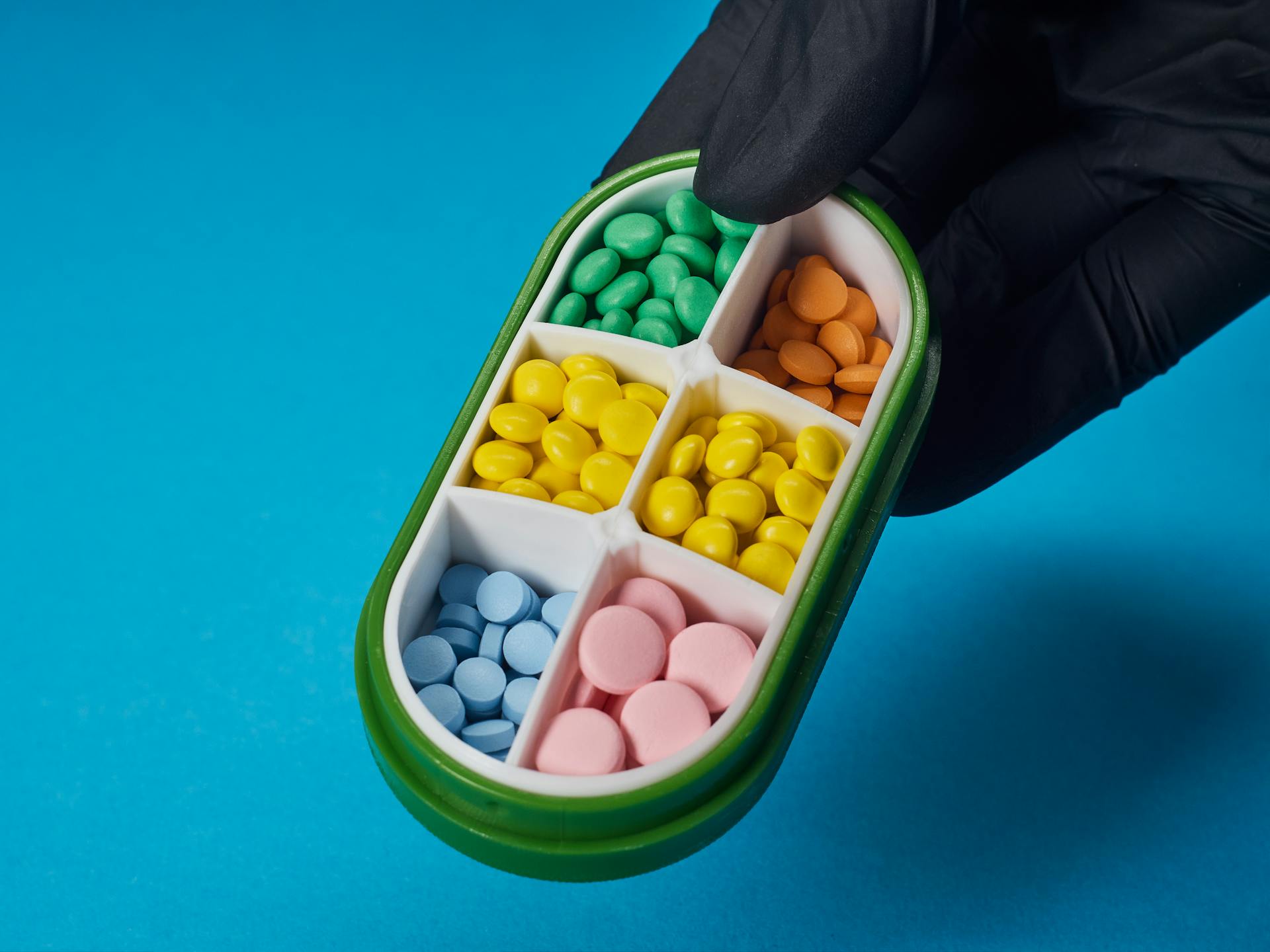 Close-up of pills in a divided container held by a gloved hand, on a blue background.