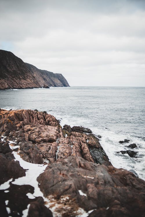 Stony shore with rough cliff washed by water of foamy ocean under cloudy gray sky