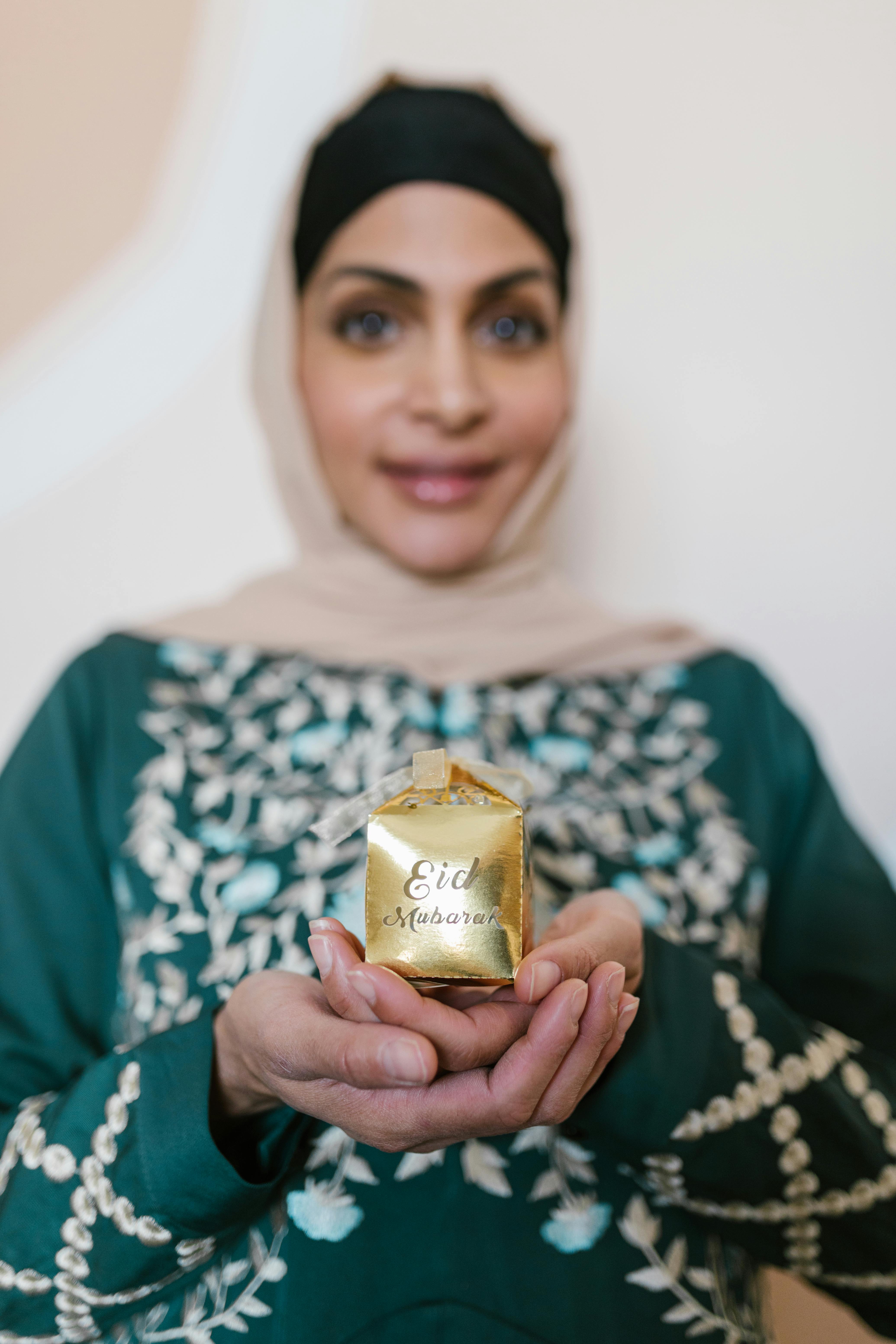 A smiling woman in traditional attire holding an Eid Mubarak gift box indoors.