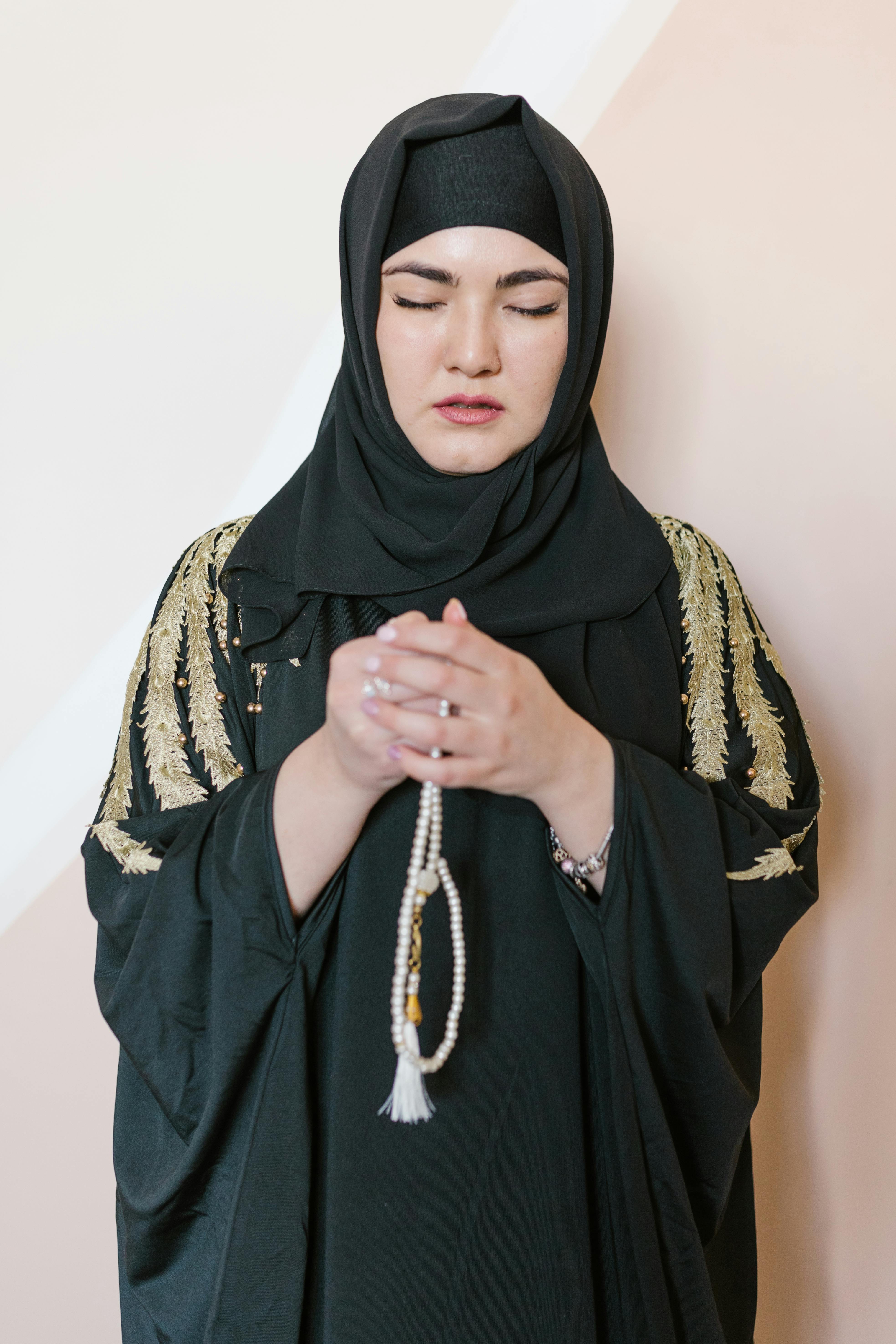 A Muslim woman in traditional dress holding prayer beads, eyes closed in deep devotion.