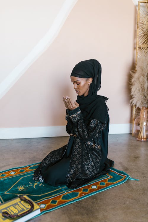 Free Woman Wearing Hijab and Traditional Dress Kneeling on a Prayer Rug Stock Photo