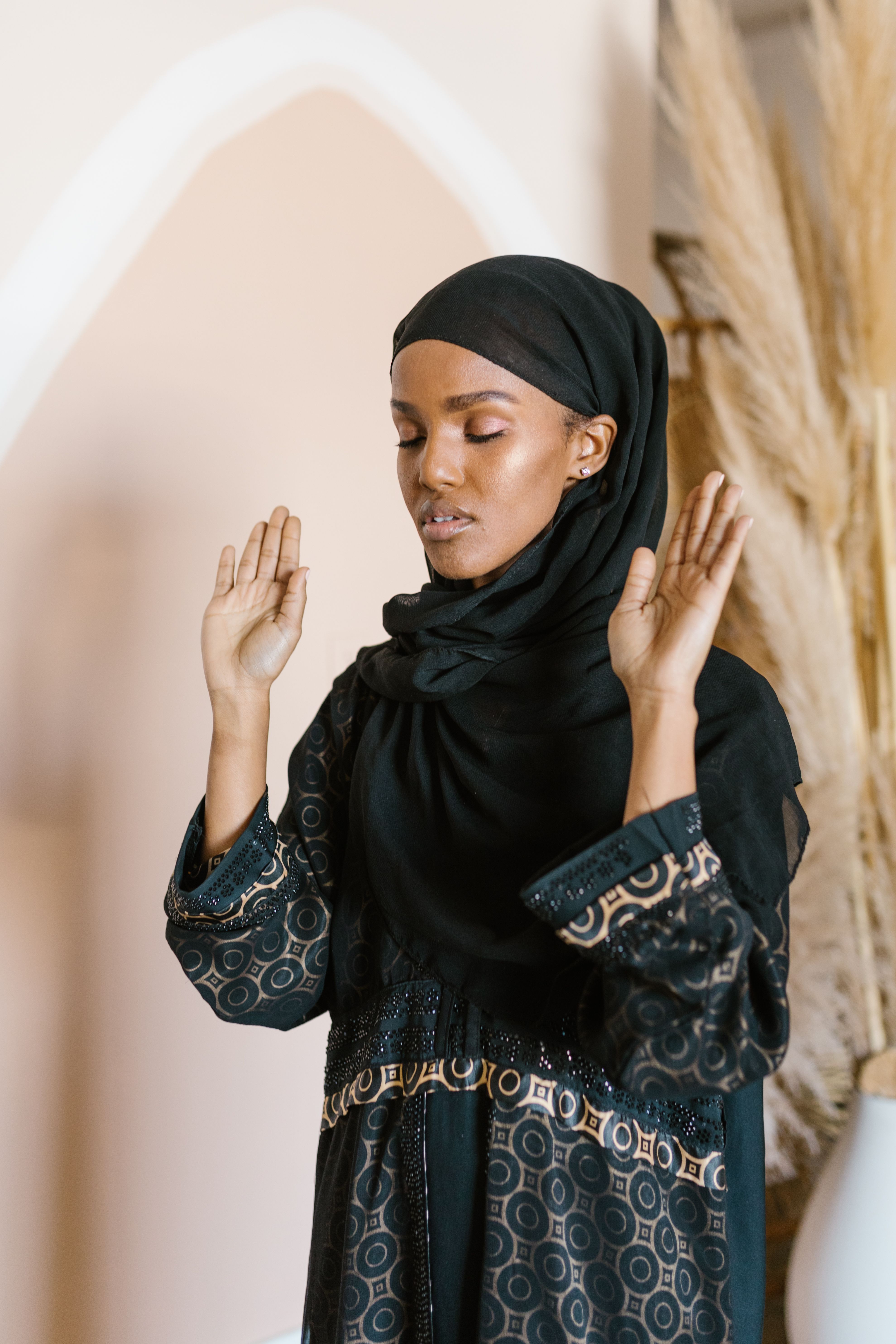 A woman in a black hijab and traditional dress praying indoors with hands raised.