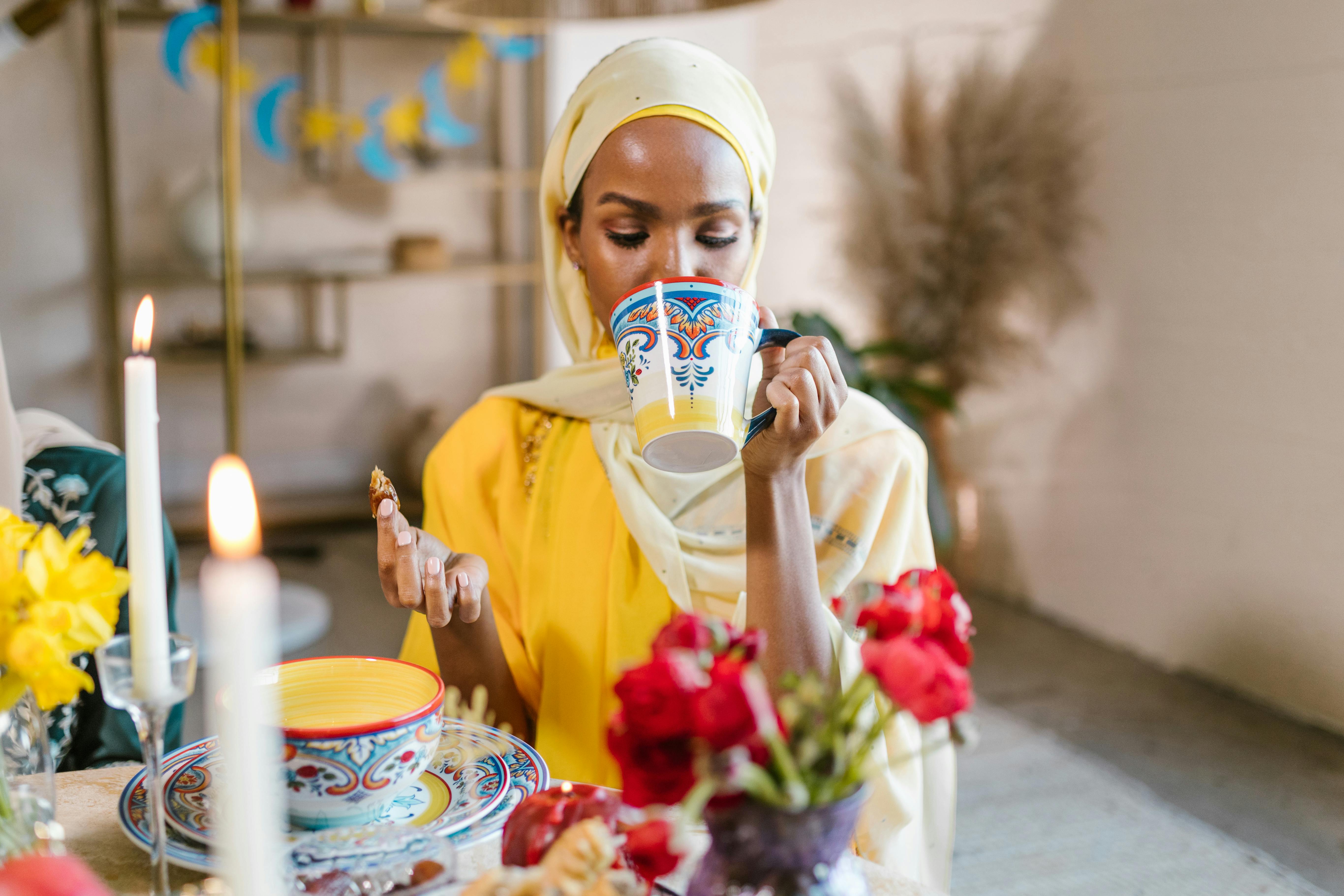 A woman in a hijab enjoys a festive meal and drink, celebrating Eid indoors.