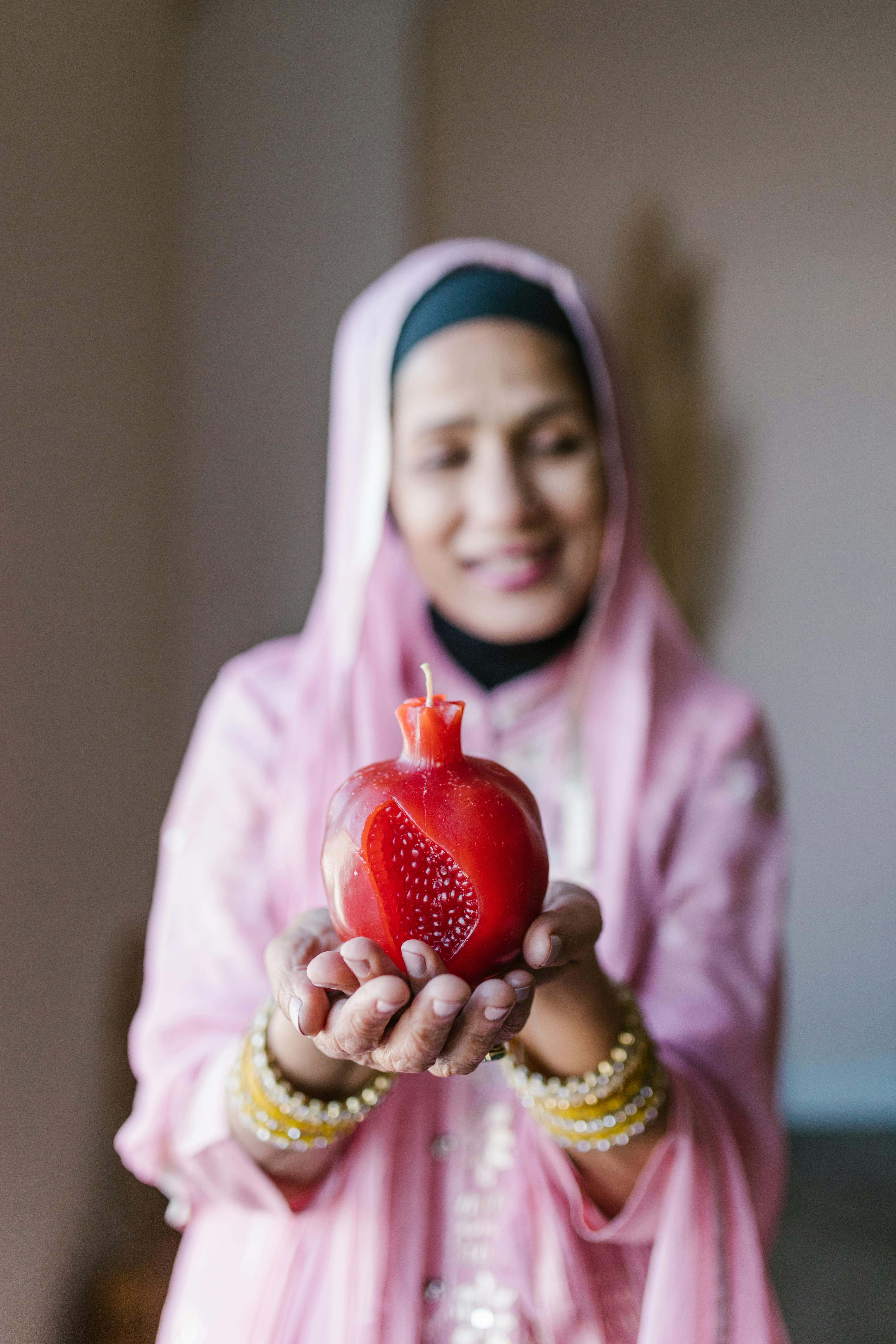 Muslim woman in hijab holding a candle shaped like a pomegranate, symbolizing warmth and festivity.