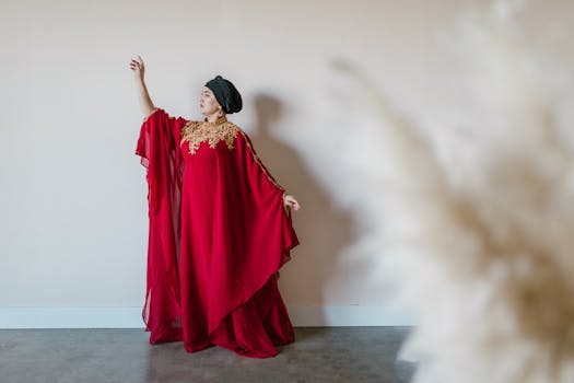 Woman in a flowing red dress with gold details posing gracefully indoors against a white wall.