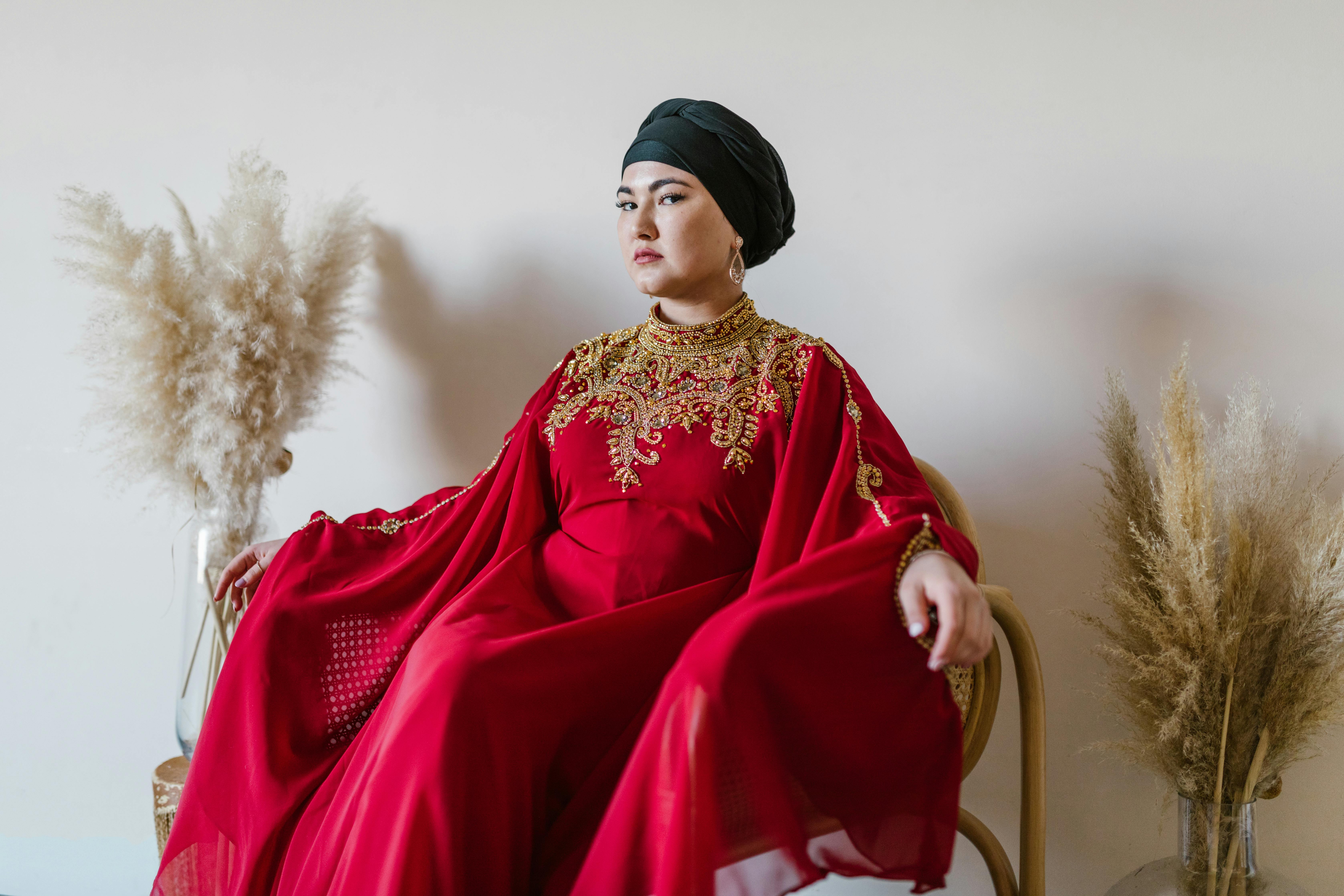 modern abayas - Portrait of a woman in a luxurious red traditional dress sitting elegantly indoors.