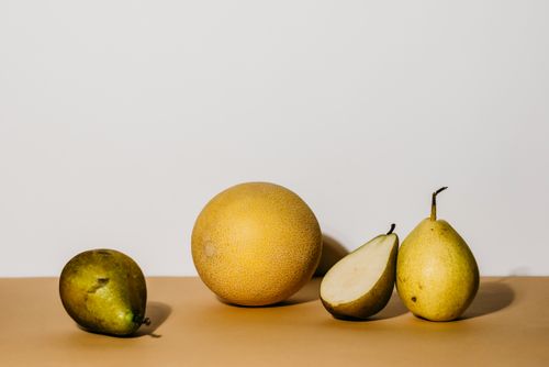 Free A minimalist still life of whole and sliced pears alongside a melon on a neutral background. Stock Photo