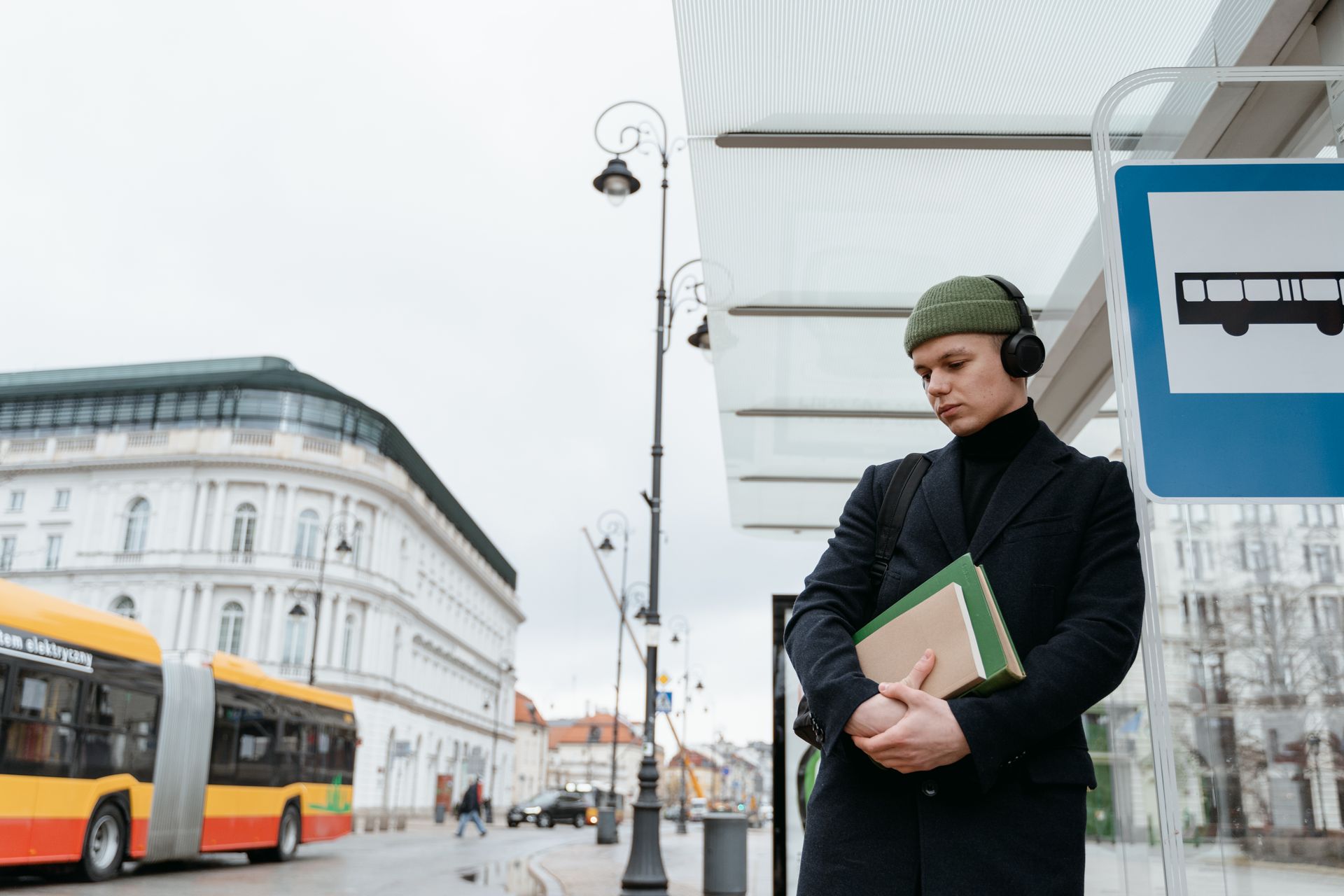 Young man in beanie and headphones waiting at a city bus stop, holding a notebook.