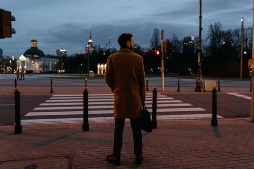 Free An adult man in a coat waits at a pedestrian crossing during a city evening. Stock Photo
