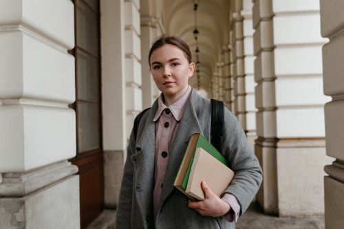 Free Young woman carrying books in an architectural hallway, dressed for fall weather. Stock Photo