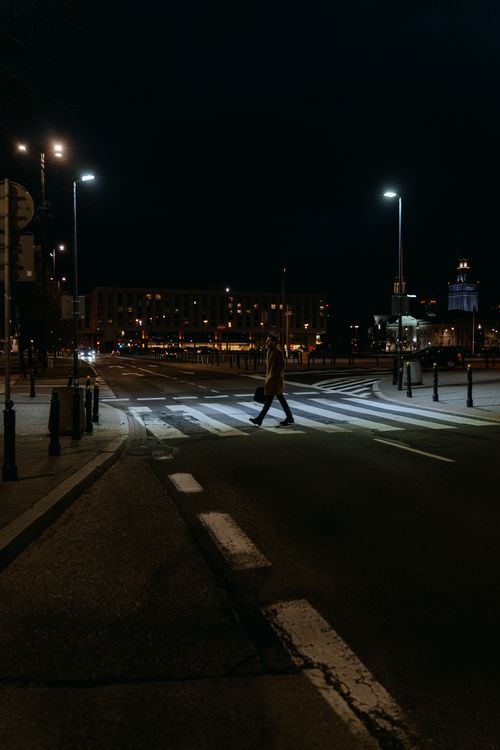 Free A solitary figure crosses a quiet, illuminated street at night in an urban setting. Stock Photo