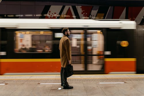 A Man in Brown Coat Standing on a Subway Platform with a  Moving Train