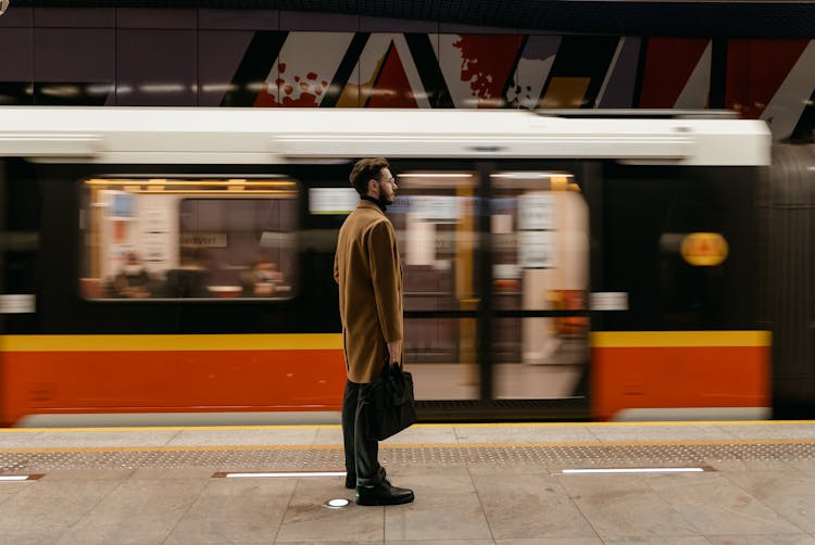 A Man In Brown Coat Standing On A Subway Platform With A  Moving Train