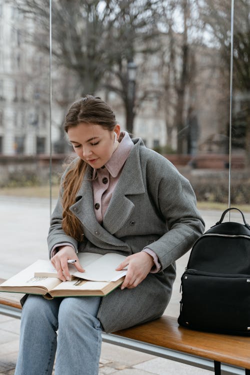 
A Woman in a Gray Coat Reading a Book while Sitting at a Bus Stop