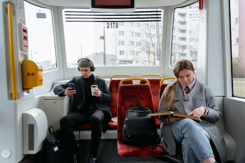 Free A Woman in Gray Coat Reading a Book while Sitting Near the Man Wearing Headphones while Holding His Mobile Phone Stock Photo