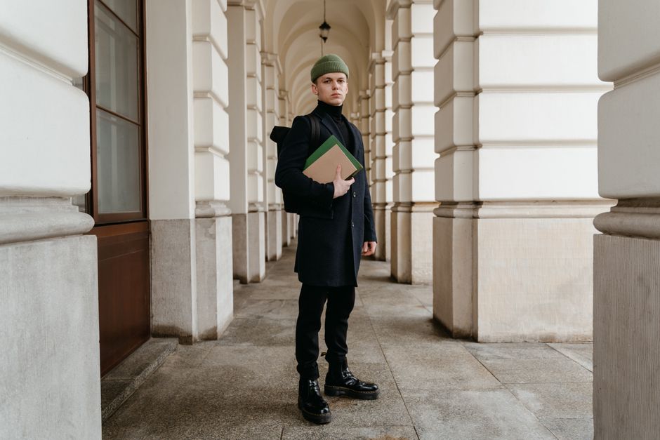 A young man in a dark coat and beanie stands in a classic archway holding books. A young man in a dark coat and beanie stands in a classic archway holding books.