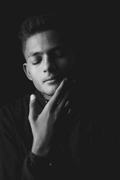 Black and white of young ethnic pensive male contemplating with eyes closed on black background