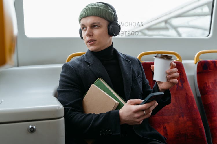 A Man In Gray Coat Sitting Inside The Bus While Holding A Cup Of Coffee And His Mobile Phone