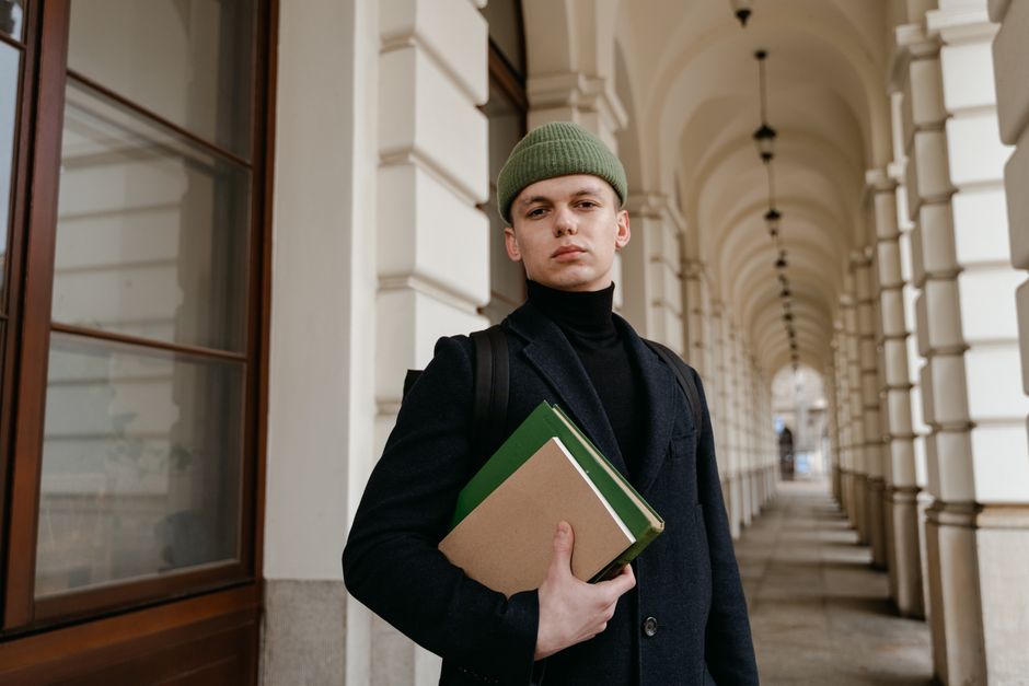 A fashionable young man in a green beanie holds books in a historic archway. A fashionable young man in a green beanie holds books in a historic archway.