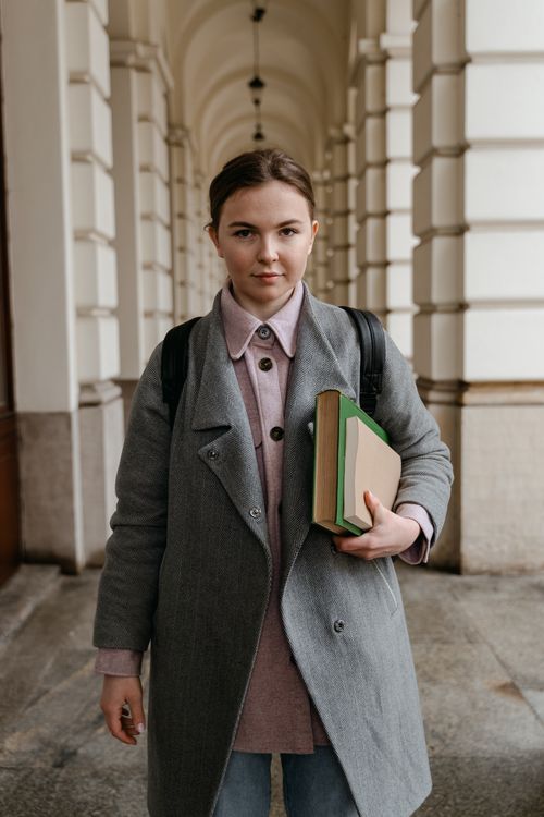 Free Young woman in a gray coat holding books in an elegant hallway with arches. Stock Photo