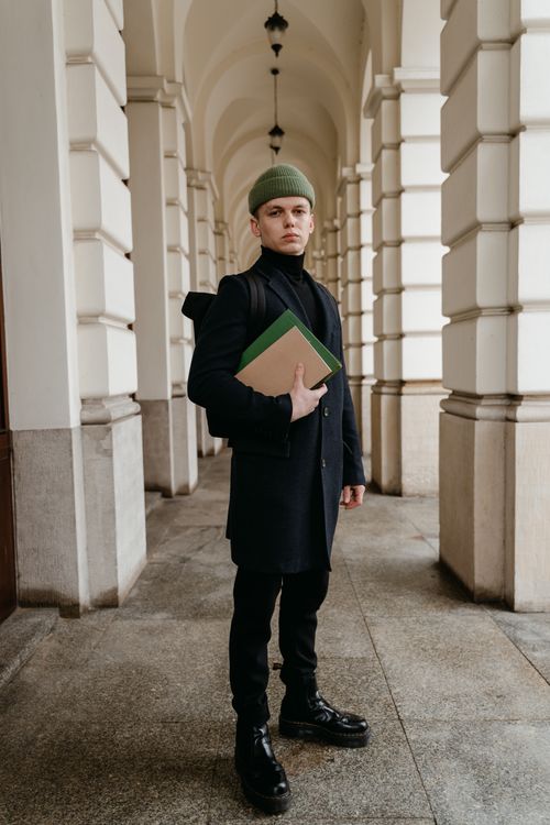 Free A fashionable young man stands in a classic hallway holding books, wearing a green beanie. Stock Photo