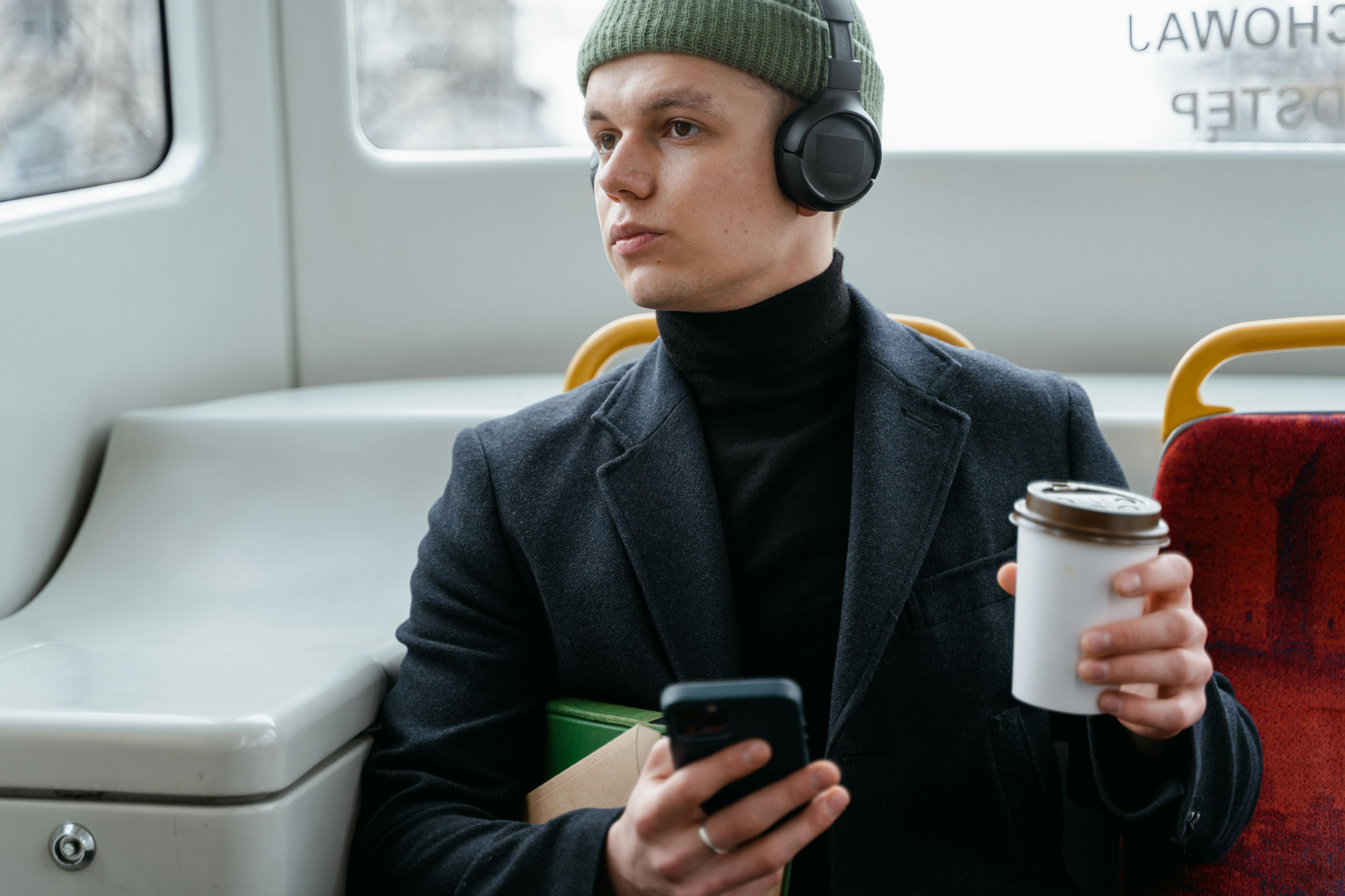 Man in urban attire with coffee and smartphone on a train, wearing headphones and a beanie.