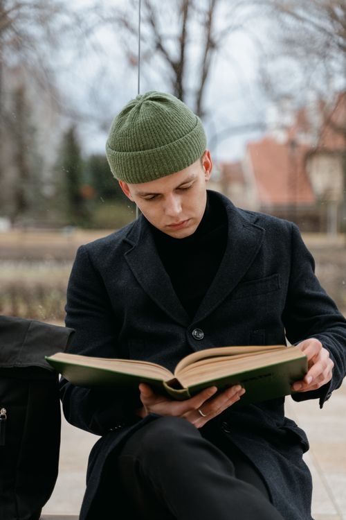 Free A man in a black coat and green knitted cap reading a book on a bench. Stock Photo