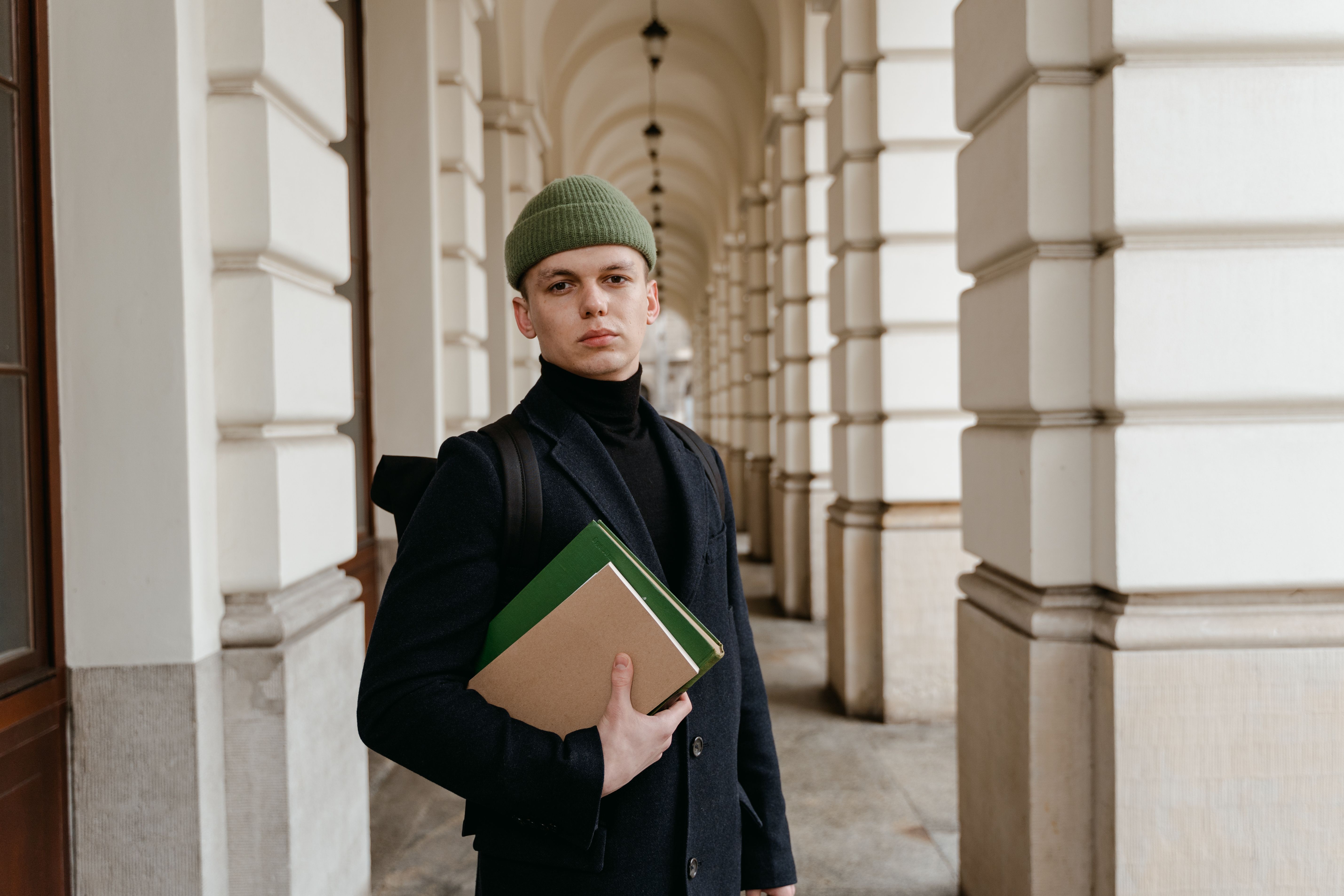 Young man in a black coat, holding books, standing in an elegant corridor.